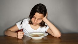Ein Mädchen sitzt betrübt vor einem Teller Suppe