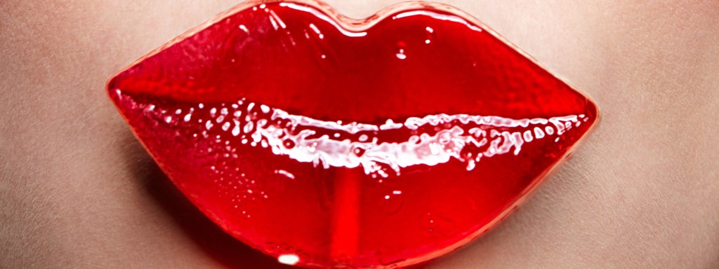 Ein roter Lolli in Form eines Kussmunde wird vor den Mund einer Frau gehalten.