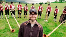 Mark Forster posiert vor einer Gruppe mit Alphornbläsern auf einer Wiese.