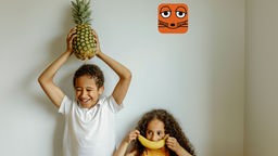 Eine Junge hält eine Ananas über den Kopf und ein Mädchen hält sich eine Banane vor den Mund.