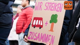 Eine Frau hält auf einer Versammlung ein Plakat in der Hand. Darauf steht "Wir streiken zusammen" und ein roter Bus und ein Baum sind mit Farbe gemalt.