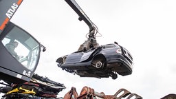 Ein Kran greift bei einem Autoverwerter ein altes Schrottauto und hebt es in eine Schrottpresse.