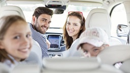 Auf dem Bild ist eine vierköpfige Familie zu sehen, die in einem Auto sitzt und in die Kamera lächelt.