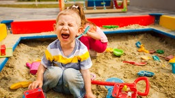 Ein Junge schreit laut, während er in einem Sandkasten sitzt. Hinter ihm sitzt ein Mädchen.