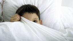 Ein Junge liegt im Bett und zieht sich vor Scham die Bettdecke über das halbe Gesicht.