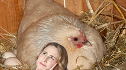 Weißes Huhn sitzt im Stroh und brütet, davor: Maus-Moderatorin Marie grübelnd