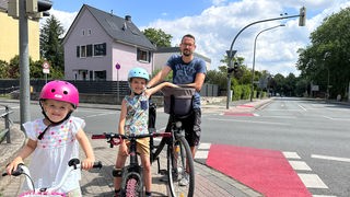 Ein Vater steht mit seinen zwei Kindern an der Kreuzung. Sie sind mit Fahrrädern unterwegs.