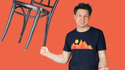 Mausmoderator André hält einen Bistrostuhl mit der rechten Hand am Stuhlbein in die Luft
