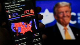 Wahlergebnis der US-Wahlen auf einem Smartphone, im Hintergrund ein Fernsehbild von Donald Trump