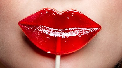 Ein roter Lolli in Form eines Kussmunde wird vor den Mund einer Frau gehalten.