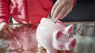 Eine Kinderhand steckt eine Euro-Münze in ein rosa Sparschwein
