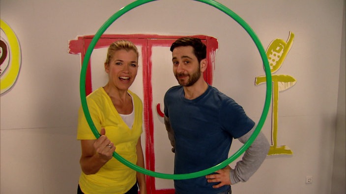 Anke Engelke und Denis Moschitto stehen nebeneinander und Anke hält einen Hula Hoop Reifen in der Hand