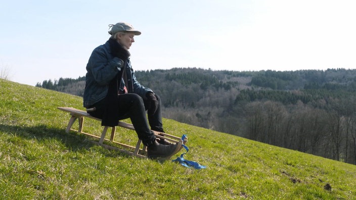 Laura sitzt auf einem Schlitten auf auf einem grünen Wiesenhang 