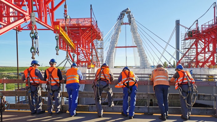 Bauarbeiter stehen mit dem Rücken zu uns und sehen nach unten auf der Brücke auf die Baustelle