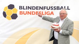 Uwe Seeler vor einem Plakat mit dem Logo und der Beschriftung der Blindenfußball-Bundesliga.