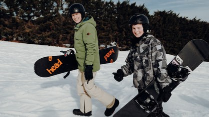 Johannes und Trainer Michel tragen ihre Snwoboards einen verschneiten Berg hoch. 