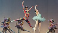 Der Tänzer Benjamin Millepied tanzt in einem Harlekin-Kostüm gemeinsam mit der Ballerina Alexandra Ansanelli das Stück „Harlequinade“. Im Hintergrund befinden sich Tänzer*innen des New York City Ballets, ebenfals in Harlekin-Kostümen.