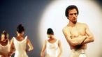 Aufnahme aus dem Jahr 1978,  auf der Rudolf Nurejew mit freiem Oberkörper, verschränkten Armen und einer weißen Balletthose zu sehen ist. Links im Hintergrund stehen drei Tänzerinnen in weißen Balletttrikots. 