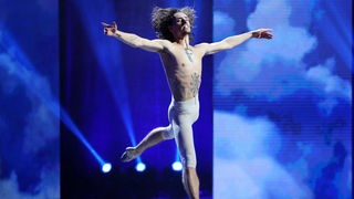 Der Tänzer Sergej Polunin mit ausgebreiteten Armen im Sprung mit nacktem Oberkörper und eines bis zu den Knien reichenden, weißen, eng anliegenden Hose vor blauem Hintergrund während einer Aufführung im Frühjahr 2020. 