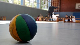 Basketball liegt auf dem Boden einer Schulturnhalle, im Hintergrund sitzen Kinder.