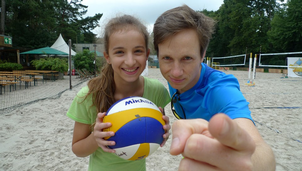 Alessia hält Volleyball, Johannes steht neben ihr und zeigt mit Finger in Richtung Kamera.