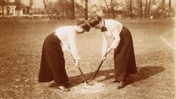 Foto von 1899 - Zwei Frauen in langen Röcken spielen Feldhockey.