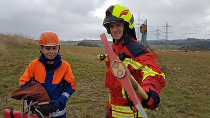 Johannes und Felix stehen in Feuerwehrausrüstung mit verschiedenen Gerätschaften auf einer Wiese.