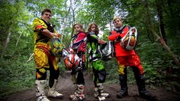 Vier Motocross-Fahrer stehen nebeneinander im Wald und gucken siegessicher in die Kamera.