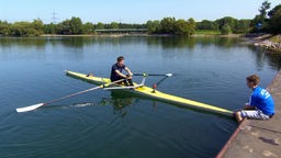 MOderator Johannes Büchs sitzt in einem Einer während Trainer Rocco das Boot auf dem Steg sitzend am Bug festhält.