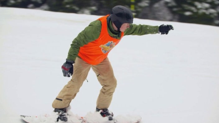 Johannes fährt auf dem Snowboard und zeigt mit einem Arm nach vorne.