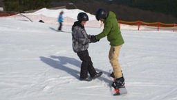 Johannes steht auf Snowboard und wird von Michel festgehalten.
