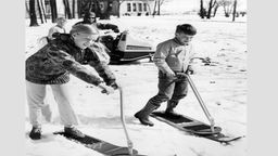 Schwarz-weiß: Zwei Kinder mit 'Ski-boards' im Schnee.