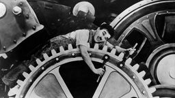Schwarz-weiß Foto aus dem Film 'Moderne Zeiten': Charlie Chaplin liegt auf riesigem Zahnrad und dreht an Schrauben.