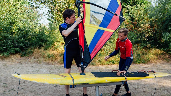 Johannes steht an Land neben seinem Surfboard, Hannes zeigt ihm, wie er das Segel halten soll.