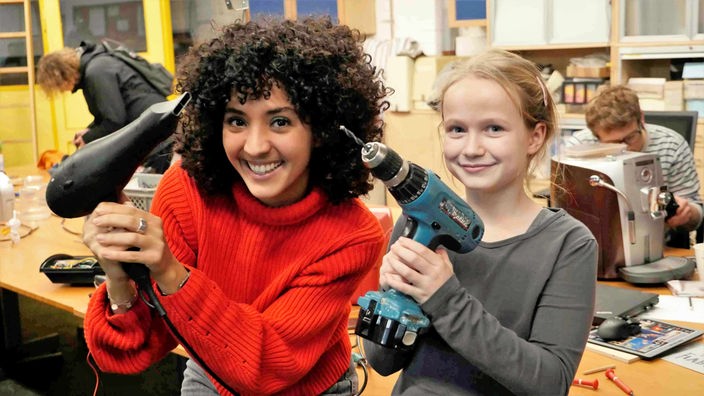 Siham mit einem Fön und ein Mädchen mit einer Bohrmaschine in einer Werkstatt