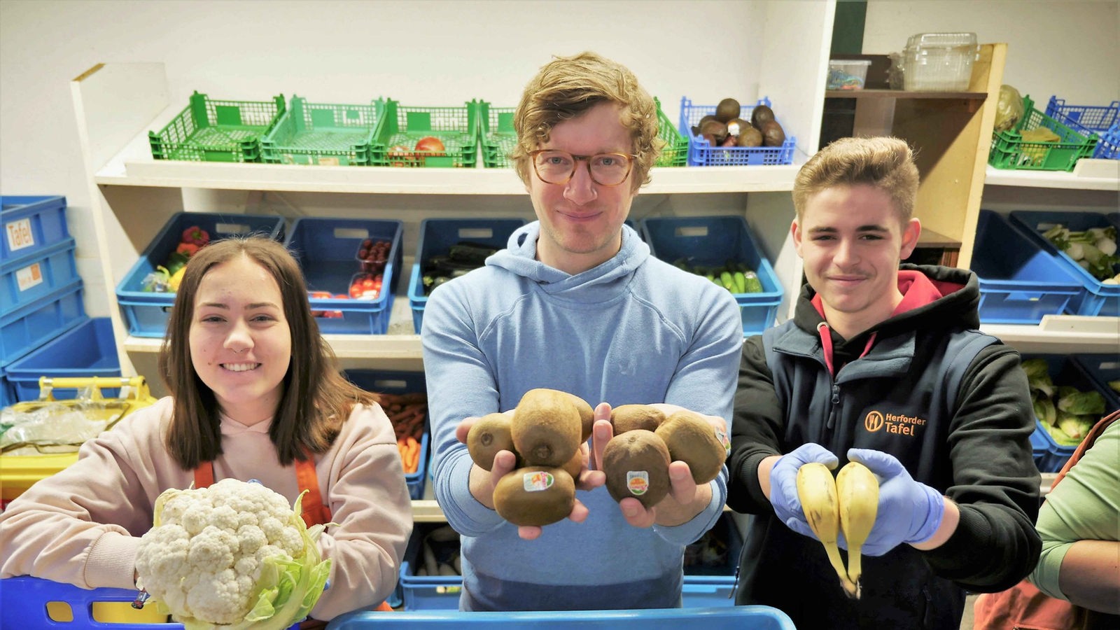 Robert, Lara und Jacq mit Obst und Gemüse in den Händen vor einem Obst- und Gemüseregal