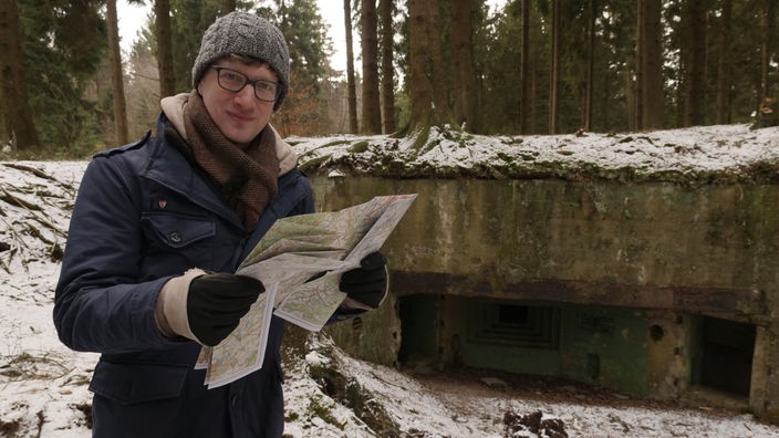 Robert steht mit Landkarte im Schnee vor einem Bunker im Wald