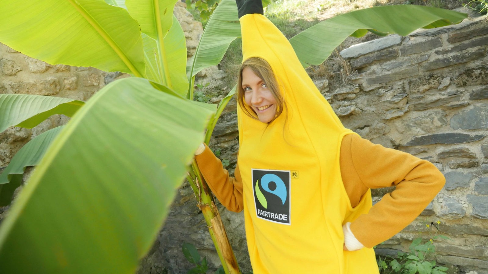 Jana im Bananenkostüm mit Fairtrade-Aufkleber vor einer Bananenstaude