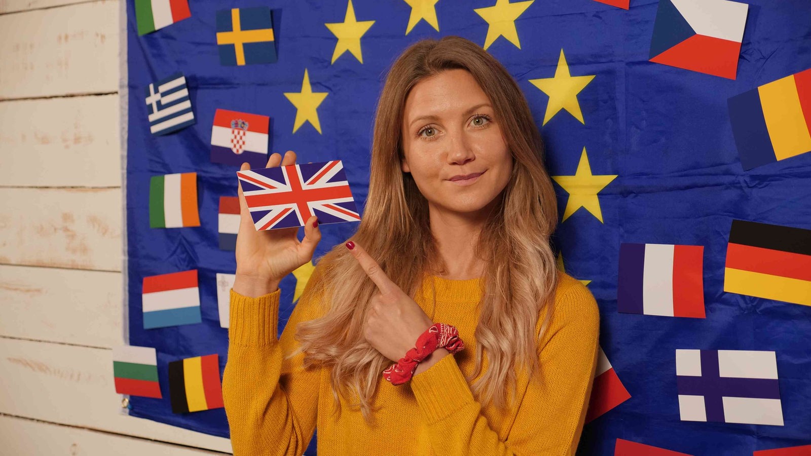 Jana vor einer weißen Holzwand mit großer EU-Flagge, in der Hand hat sie die Nationalflagge des Vereinigten Königreichs Großbritannien und Nordirland.