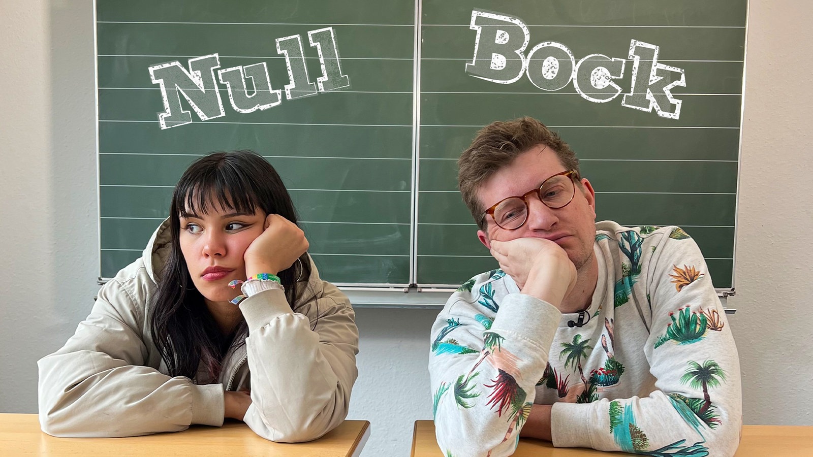 Robert sitzt mit Protagonistin Izabel in einem Klassenraum. Ihre Blicke sind lustlos und genervt. Auf der Tafel hinter ihnen ist „Null Bock“ zu lesen.