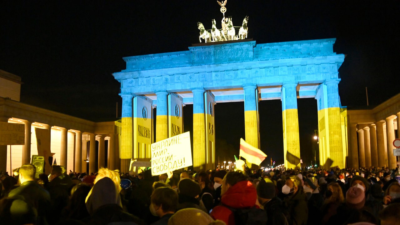 Demonstrierende vor dem Brandenburger Tor, das in den Farben der ukrainischen Flagge erstahlt. 