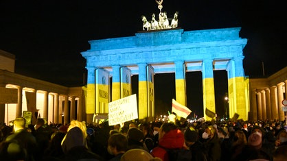 Demonstrierende vor dem Brandenburger Tor, das in den Farben der ukrainischen Flagge erstahlt. 