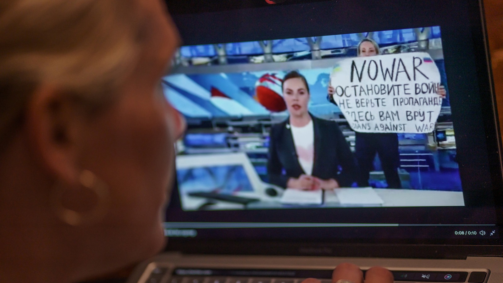Auf einem Laptop-Monitor sieht man eine russische Nachrichtensprecherin hinter der eine Frau mit einem Schild steht, Aufschrift: No War.