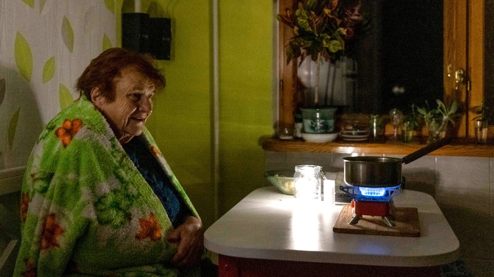 Eine ältere Frau hat sich in eine Decke gehüllt und sitzt in der Dunkelheit in ihrer Wohnung vor einem kleinen Gaskocher.