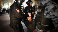 Ein russischer Demonstrant, der von Polizisten weggetragen wird, hält einen Ausweis hoch.