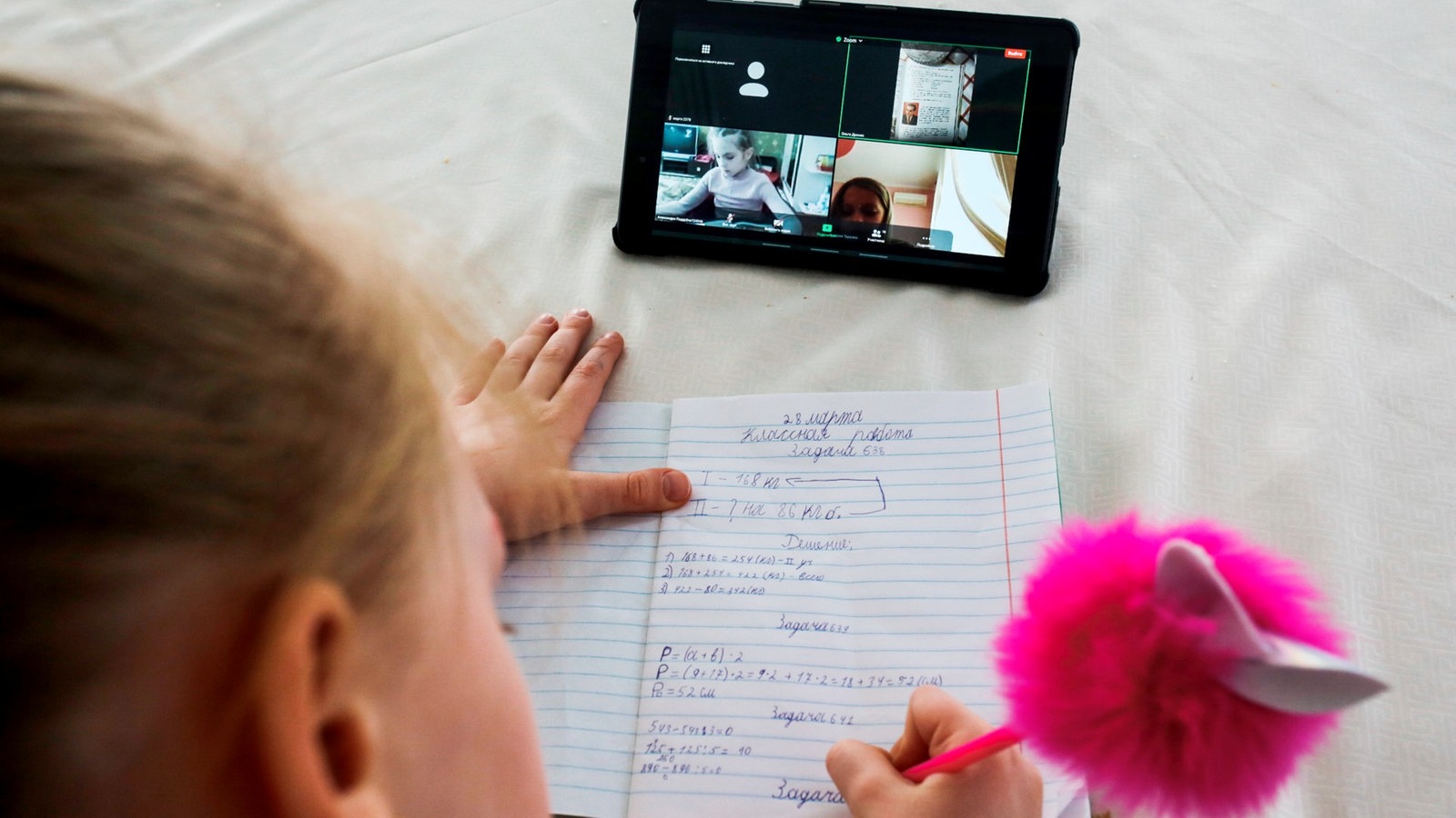 Ein Mädchen schreibt in Schulheft, vor ihr liegt ein Tablet, auf dem andere Kinder zu sehen sind.