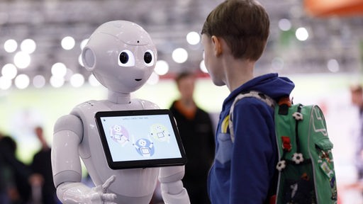 Ein Junge steht vor dem Roboter Pepper, der ein Tablet um den Hals hängen hat.
