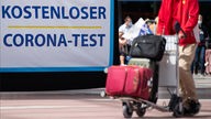 Kofferwagen mit mehreren Gepäckstücken vor einem Plakat mit der Aufschrift: Kostenloser Corona-Test.