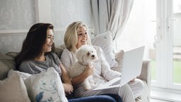 Zwei Frauen sitzen mit Hund und Computer auf einem Sofa.