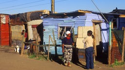 Zwei schwarze Frauen stehen vor einer aus Brettern zusammengezimmerten Hütte.